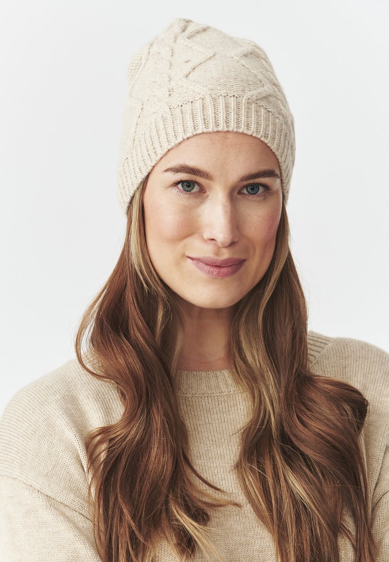 Beige kootud beanie koos tekstuuriga kaabelmustriga, pehme materjal ja sooniline äär. Kanda koos sobiva heleda beeži sviitriga.