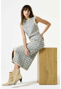 Vrouw zit op een houten blok en draagt een mouwloze blauw en beige tweed jurk met een voorknoop en beige suède enkellaarsjes.