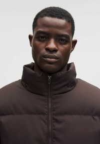 Ruskea pehmeä takki, jossa on korkea kaulus, etuvetoketju ja napitusyksityiskohdat. Sujuva pinta, yhtenäinen väri ja minimalistinen muotoilu.