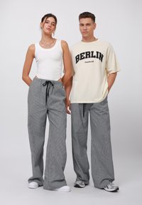 Grå rutiga vida byxor med svart dragsko, tillsammans med en vit linne och en krämfärgad t-shirt med fet svart text "BERLIN".