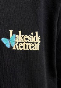 Čierna látka s krémovým textom "Lakeside Retreat" a modrým motýlikom, ktorý čiastočne prekrýva písmeno L.