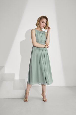 Woman in sleeveless, pleated light green midi dress and gold heels posing with one hand touching her hair against plain white background.