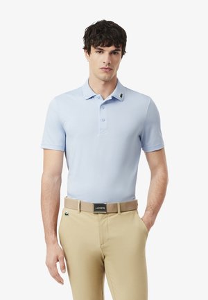 Man draagt lichtblauw poloshirt met korte mouwen en beige broek met bijbehorende beige riem tegen een effen witte achtergrond.