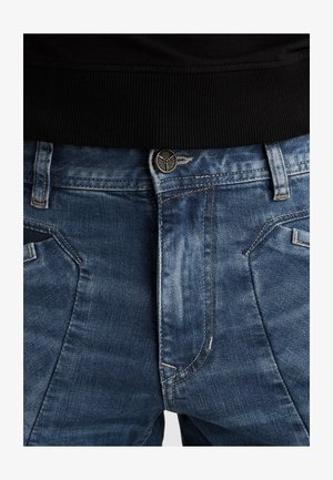 Gros plan sur le devant d'un jean en denim bleu avec bouton, braguette zippée, passants de ceinture et coutures contrastées, porté avec un haut à taille côtelée noire.