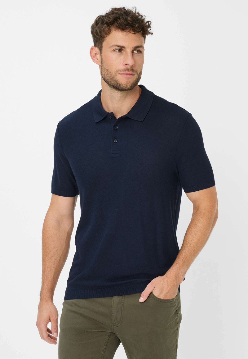 Marineblaues Poloshirt mit kurzen Ärmeln, Drei-Knopf-Leiste und strukturiertem Stoff. Legerer Schnitt kombiniert mit hellgrünen Hosen.