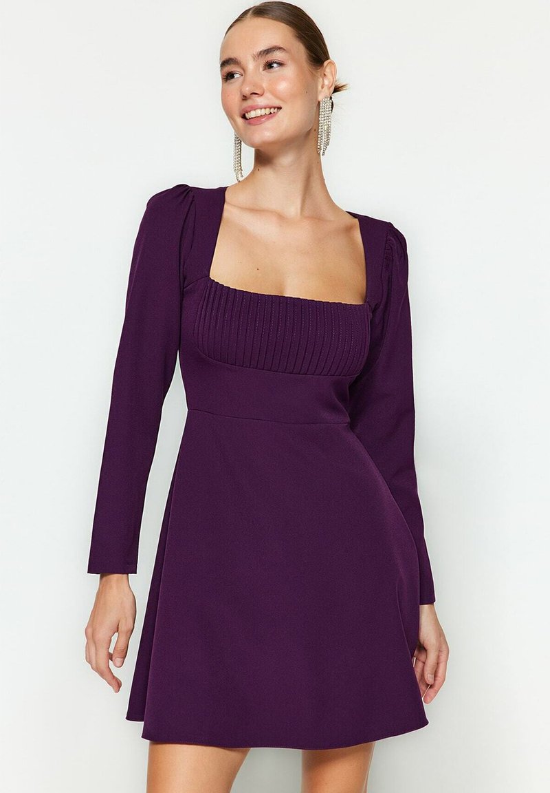 Robe violette à manches longues, décolleté carré et corsage côtelé. La jupe s'évase en dessous de la taille, créant une silhouette ajustée.