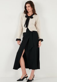 Cream sweater with black lace bow and cuffs, paired with a black wrap skirt featuring a side slit, accented with a metallic detail. Black heels.