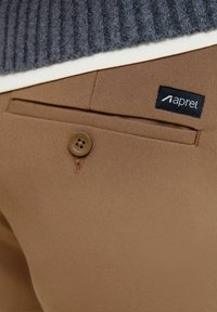Brown fabric trousers with a back pocket, featuring a button and a black label with white text. Textured gray knit sweater visible above.
