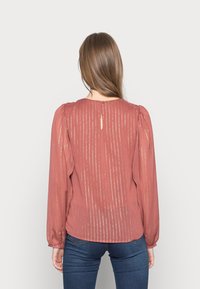 Blusa de color óxido con mangas largas abullonadas, rayas doradas verticales, una abertura en forma de ojal en la espalda y una textura ligera y fluida.