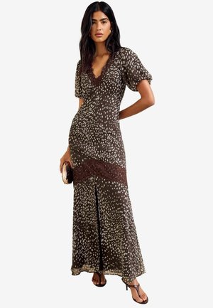 Woman wearing a brown, patterned maxi dress with puff sleeves and lace detail, holding a small clutch and wearing heeled sandals.