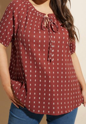 Femme portant un chemisier rouge rouille à motifs carrés blancs, manches courtes, avec un détail à nouer au niveau du col, associé à un jean bleu.