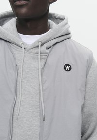 Grijze mouwloze jas over een grijze hoodie, met een ritsluiting, trekkoorden en een ronde zwarte patch met een wit logo op de borst.