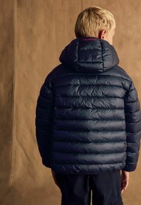 Chaqueta acolchada en color navy con diseño de rombos horizontales y capucha. Presenta un logo en el cuello posterior y una textura suave y brillante.
