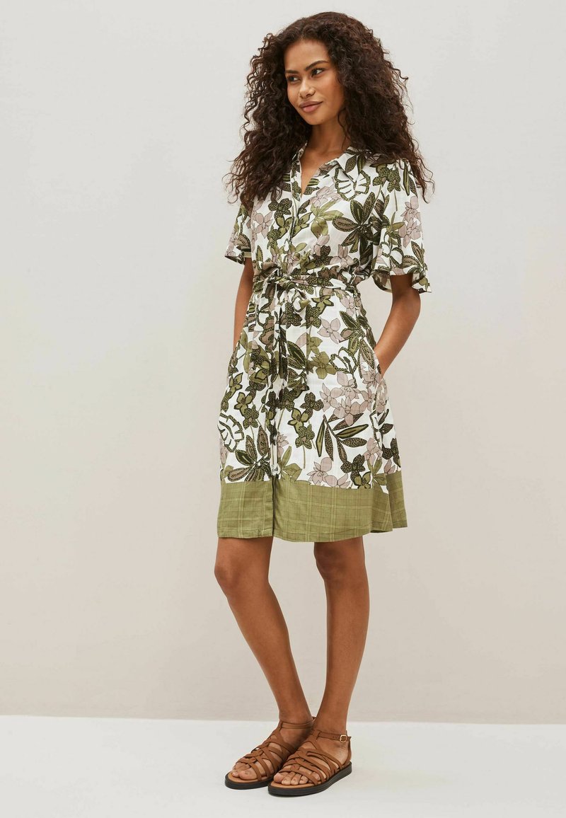Floral kurzes Kleid mit grünen und weißen Mustern, Knopfverschluss vorne, kurzen Ärmeln, tailliertem Bund und strukturiertem grünen Saum, kombiniert mit braunen Sandalen.