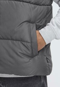 Grå puffer vest med en glatt, quiltet tekstur og sidelommer. Har et logomerke på kanten og er laget over en grå langermet skjorte.