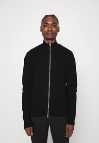 Sudadera negra con cremallera y cuello alto, textura acanalada, corte ajustado, cremallera plateada y mangas largas, combinada con pantalones oscuros.
