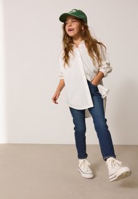 Camicia bianca a bottoni, jeans blu e sneakers bianche. Cappellino verde con testo. Capelli sciolti, in una posa casual in piedi. Sfondo semplice.