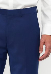 Pantalones ajustados azul marino con una textura suave, frente plano y lazos para el cinturón. Se pueden ver bolsillos laterales y una silueta ajustada.