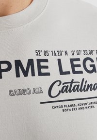 Sudadera gris claro con texto en azul marino. El texto incluye coordenadas geográficas y detalles de la marca. Cuello acanalado y textura de tela suave.