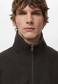 Tmavohnedá bunda na zips s vysokým golierom, s hladkou textúrou a kovovým zipsom. Jednoduchý dizajn, bez výrazných vzorov alebo akcentov.