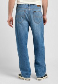 Homme portant un jean en denim bleu droit et des baskets noires, debout avec les mains détendues le long du corps, vue de dos.