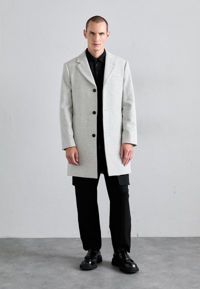 Light gray wool coat with a notched collar, single-breasted front, and black buttons. Worn over a black shirt and paired with black pants.