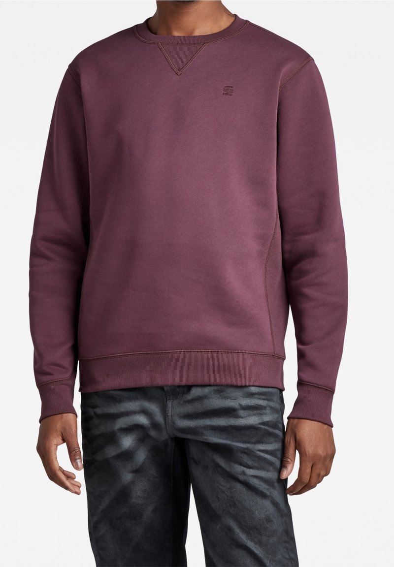 Bordowy sweatshirt z okrągłym dekoltem i ściągaczami. Posiada subtelne logo na piersi oraz teksturowane szczegóły szwów. Połączony z ciemnymi spodniami.