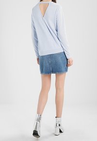 Pull long bleu clair avec une découpe au dos, associé à une jupe en denim et des bottines à talons carrés argentés. Texture de tissu lisse.