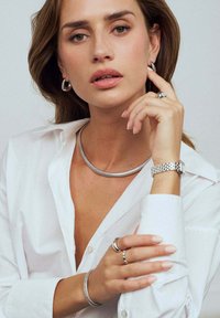 Smycken i silver inkluderar en tjock, flexibel halskedja, örhängen i form av hoops, en stilren klocka och flera ringar. Presenteras med en krispig vit skjorta.