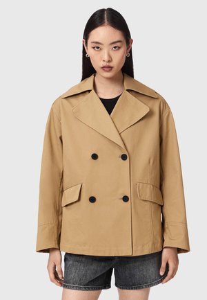 Kvinde iført en beige dobbeltradet jakke med sorte knapper og klaplommer, kombineret med mørke cowboyshorts, stående mod en ensfarvet baggrund.