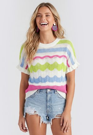 Vrouw die lacht en een meerkleurig gestreept gebreid topje draagt met gerimpelde mouwen en versleten denim shorts.