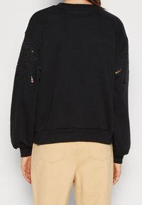 Felpa nera con maniche lunghe e a sbuffo, caratterizzata da dettagli con tagli sulle spalle. Tessuto morbido e orlo a coste, indossata con pantaloni beige.