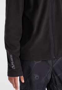 Nahaufnahme einer Person, die eine schwarze Polartec-Fleecejacke und eine dunkle, gemusterte Hose trägt, wobei die Jacke teilweise zugezippt ist.