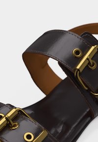 Σκούρο καφέ δερμάτινο σανδάλι με χρυσά μεταλλικά αγκράφες και οπές σε δύο πλαίες λουρίδες, σε λευκό φόντο.