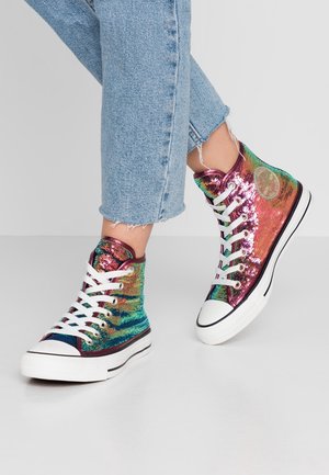 Feet wearing blue denim jeans and high-top sneakers covered in shiny multicolored sequins with white laces and rubber soles.