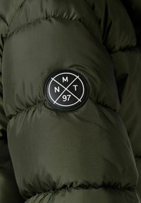 Manica di giacca puffer verde oliva con tessuto testurizzato, un patch rotondo nero con lettering bianco e sezioni trapuntate per l'isolamento.