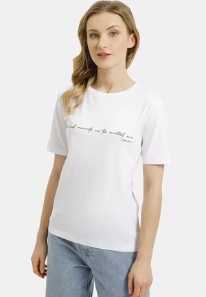 Kobieta o jasnych włosach, ubrana w białą koszulkę z napisem na piersi oraz jasnoniebieskie dżinsy, stojąca na jednolitym tle.