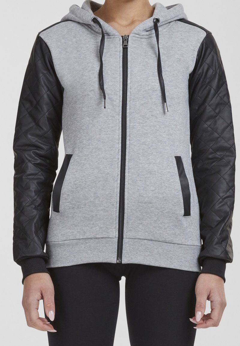 Sweat à capuche gris zippé avec manches matelassées noires, bordure de poche noire et cordons noirs, porté par une personne debout, les bras détendus.