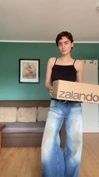 A kezében tartott "zalando" felirattal ellátott kartondoboz, zöld fal előtt; fekete topban és világoskék, bő szárú farmernadrágban van.