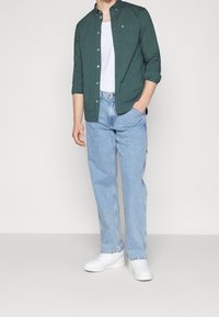 Man in een donkergroene knoopshirt over een wit t-shirt, lichte blauwe jeans en witte sneakers, staand met één hand in de zak.