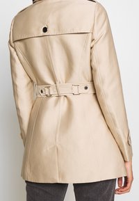 Beige pásek s klopovým límcem, knoflíkovými detaily a zdobením na zadní straně. Vyrobeno z hladkého materiálu s přiléhavým střihem.