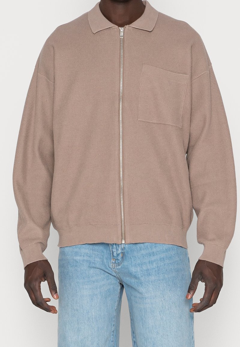 Maglione lavorato a maglia beige con zip e taschino sul petto, indossato con jeans di denim azzurro chiaro.