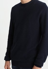 Mörkblå stickad tröja med texturerat mönster, rundad halsringning och ribbade ärmar. Tyget verkar tjockt och varmt.