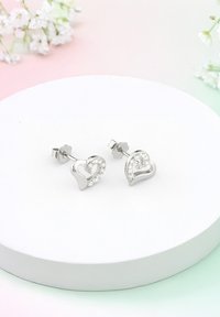 Silverfärgade hjärtformade örhängen med en glittrande yta av inbäddade klara stenar, placerade på en slät rund visningsbas.