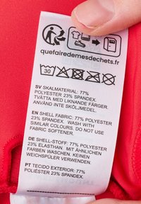 Punainen kangasvaroitusmerkki, joka näyttää pesuohjeet. Materiaalikoostumus: 77 % polyesteriä, 23 % spandexia. Monikielinen teksti. Sisältää kierrätysikonin.