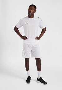 Conjunto branco de camisa atlética e calções feito de tecido leve, com riscas laterais pretas e logótipo, combinado com sapatilhas atléticas pretas e meias brancas.