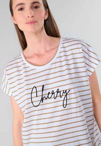 Camisa blanca con rayas doradas horizontales, cuello redondo y mangas cortas. Presenta la palabra "Cherry" en текстом cursivo negro.