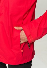 Veste imperméable rouge à texture lisse ; avec une main dans une poche latérale, un poignet en Velcro et un cordon de serrage à l'ourlet.
