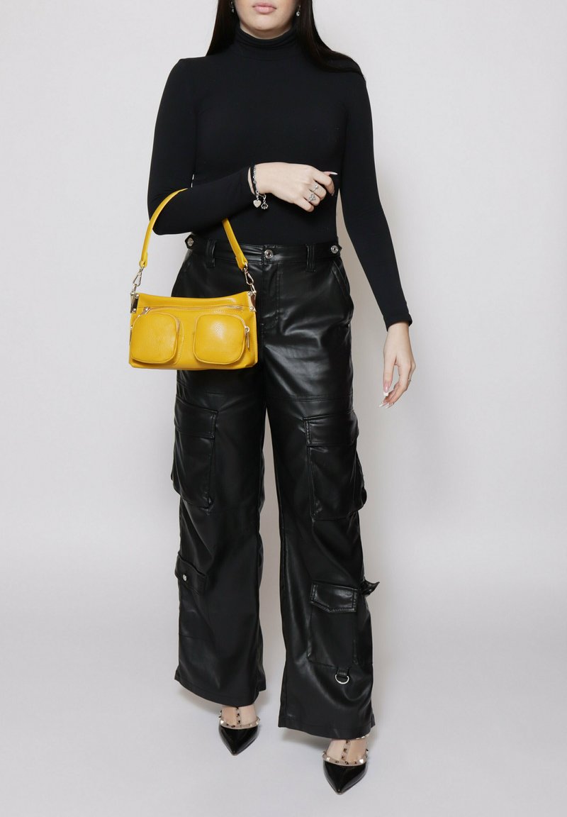 Maglione a collo alto nero a maniche lunghe, pantaloni cargo in pelle nera oversize con tasche, e una piccola borsa in pelle gialla con dettagli in zip.