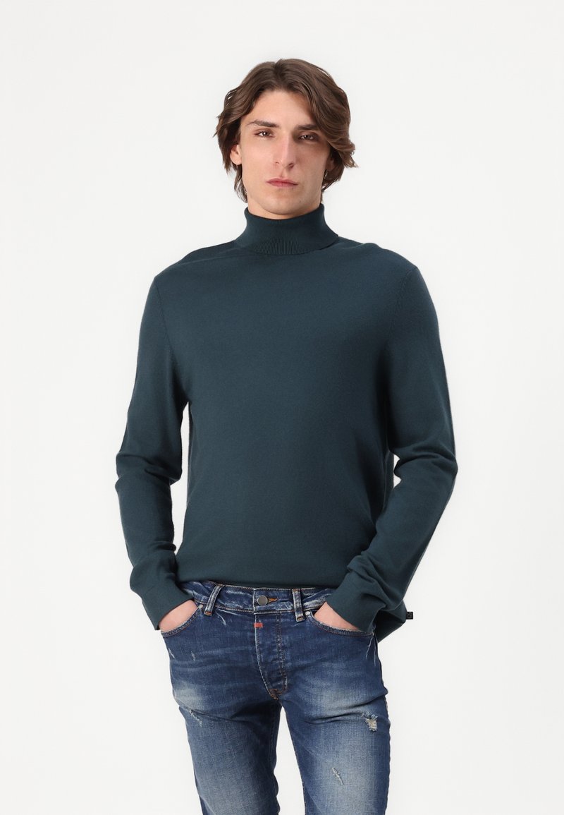 Pull à col roulé en bleu sarcelle, en tricot, avec des manches longues et une coupe ajustée, assorti à un jean bleu déchiré.